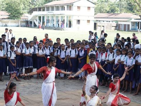 Girls in red and white saris performing a classical dance while the school watches